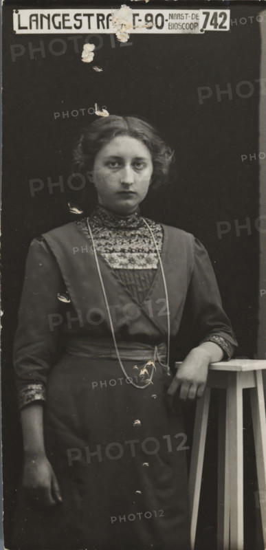 Rapid photographs, snelfoto, photographer: American Tip Top Foto Company, Alkmaar, before 1913-07-08 - after 1917-07, paper, gelatin silver print, developing gelatin silver print, height 77 mm x width 37 mm, A monochrome portrait of a young woman standing against a dark backdrop, shown from head to below the waist, with short wavy hair and a neutral facial expression; she wears a high-collared dress with a patterned yoke and decorative trim at the cuffs, a long thin necklace draped over the front of the dress, and a belt at the waist; her right arm hangs by her side while her left arm rests on a tall wooden stool with visible slatted legs, and there are small areas of surface damage or peeling visible across the photograph, including near the top and over the woman's torso and lower dress; a rectangular sign with block lettering is positioned above her head at the top edge of the image.