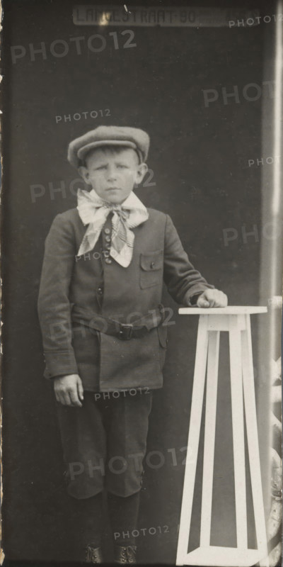 Rapid photographs, snelfoto, photographer: American Tip Top Foto Company, Alkmaar, before 1913-07-08 - after 1917-07, paper, gelatin silver print, developing gelatin silver print, height 85 mm x width 41 mm, A monochrome portrait of a boy standing against a dark backdrop, wearing a flat cap and a jacket with a wide patterned scarf tied at the neck, a belt around his waist, knee-length trousers, long socks, and lace-up boots; his left hand rests on a tall, light-colored, geometric wooden stand with vertical slats while his right arm hangs by his side, and faint scratches and wear are visible on the photograph surface.