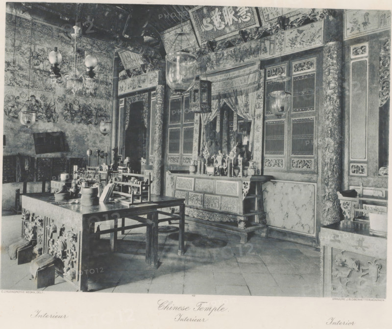 Interior of a Chinese temple, presumably in Kloempang on Sumatra, Kloempang. Chinese Temple, photomechanical print, Part of Album with heliogravures of Sumatra's east coast., number: '12', photographer: Carl J. Kleingrothe, Sumatra, cliché maker: Johann Baptist Obernetter, Munich, in or before 1898, Carl J. Kleingrothe, Johann Baptist Obernetter, photographer, cliché maker, Bavaria, paper, photoaquatint, heliogravure, height 202 mm x width 270 mm, temple, shrine, Hinduism, Buddhism, Jainism, A room filled with ornate wooden furniture and carved panels, showing a large table in the foreground with various vessels and small objects arranged on it, surrounded by wooden benches and chairs. The walls and pillars are covered with intricate painted panels and decorative reliefs, and several hanging glass lanterns and a chandelier descend from the ceiling. Behind the table an elaborately carved cabinet or altar stands against the wall, topped with vases and sculptural ornaments and flanked by ...