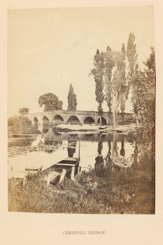 View of the Chertsey Bridge over the Thames, Chertsey Bridge, photograph, page, photographer: attributed to Francis Frith, Surrey, c. 1862 - in or before 1867, Attributed to, paper, albumen print, height 100 mm x width 140 mm, river, Thames, A sepia-toned photograph shows a stone arch bridge spanning a calm river, with multiple rounded arches and a low parapet, tall trees lining the far riverbank rising above the bridge, and reflections of the bridge and trees mirrored in the water; in the foreground there is grassy riverbank with wooden boats moored at the edge, some partially in the water and some alongside simple wooden planks, and water lilies and reeds float near the near shore.