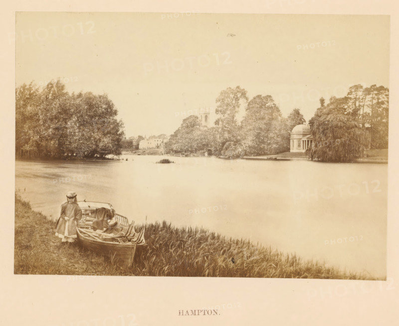 Hampton, View across the Thames at Hamton in London, photograph, page, photographer: attributed to Francis Frith, c. 1862 - in or before 1867, Richmond upon Thames, Attributed to, paper, albumen print, height 100 mm x width 140 mm, river, Thames, A sepia-toned riverside scene with tall grasses along the near bank, a small rowboat pulled up at the water’s edge, and two figures standing beside the boat wearing long coats and hats; the calm river stretches across the image toward the far bank where dense trees and a domed classical pavilion with columns sit among foliage, and beyond the trees a church tower and a cluster of buildings are visible under a pale sky.