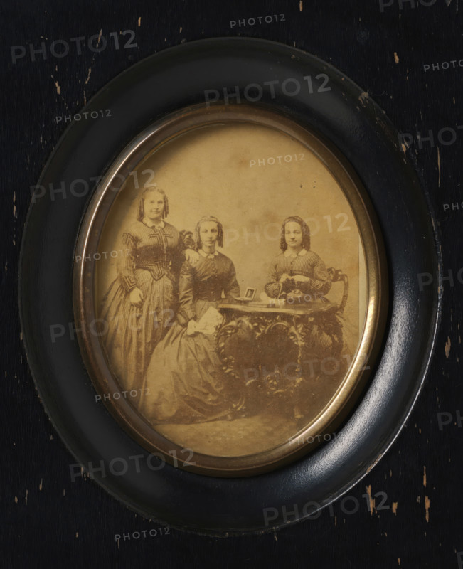 Group portrait of three unknown women around a table, with photos on top, frame, photograph, label: 'Firma Gerard Pisters, (H. Grevers), Steenweg 4, Roermond', frame maker: Firma Gerard Pisters, Roermond (Limburg), c. 1855 - c. 1860, photographer, framemaker, plant material, pasteboard, biological material, inorganic material, imitative material, paper, glass, metal, wood (plant material), imitation leather, albumen print, albumen print in frame, height 74 mm x width 60 mm, height 142 mm x width 120 mm, historical persons not known by name - BB - woman, historical persons depicted in a group, in a group portrait, An oval sepia photograph set in a round dark wooden frame with a slightly worn surface showing small chips and scratches; inside the photograph three young women in period dresses posed together, one seated at a small ornate table with a decorative carved surface, one seated beside her holding a folded handkerchief, and one standing with a hand resting on the seated woman's sh...