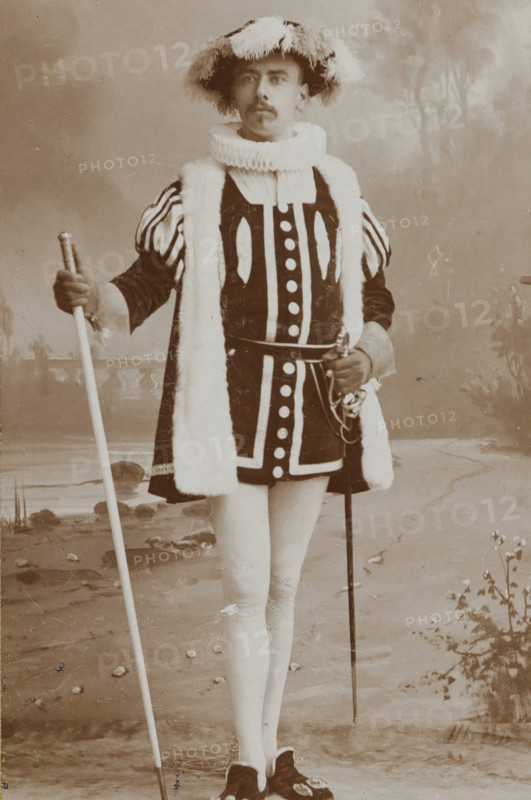 Portrait of Arnold Cornelis Maas Geesteranus in costume, album page, photograph, carte-de-visite, This photo is part of an album., name: 'AC Maas Geesteranus., 23.', inscription: 'Adolphe Zimmermans, de Riemerstraat 21., La Haye., Specialité pour les agrandissements, au charbon inaltérable.', photographer: Adolphe Zimmermans, The Hague, c. 1887, paper, pasteboard, daylight collodion silver print, height 83 mm x width 52 mm, Person, Arnold Cornelis Maas Geesteranus, historical persons, historical costume, A sepia portrait of a person standing full length in a theatrical costume, wearing a wide brimmed hat trimmed with feathers, a ruffled high collar, a dark tunic with bold contrasting trim and round decorative elements down the front, padded striped sleeves, and a long fur-trimmed stole draped over the shoulders; the person holds a long staff in one hand and a sword with an ornate hilt at the waist in the other, and wears light-colored stockings and dark shoes with decorative buckles, s...