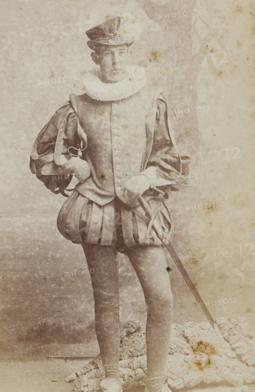 Portrait of a young man in costume, identified as HM Hijmans, album page, photograph, carte-de-visite, This photo is part of an album., inscription: 'J. Goedeljee, Photographer, HRH PRINCE ALEXANDER of the Netherlands, PRINCE of ORANGE, and HRH PRINCESS HENDRIK of the Netherlands, Hoogewoerd 160, near the Plantage.', name: 'HM Hijmans, 53.', photographer: Jan Goedeljee, Leiden, 1860 - 1905, paper, pasteboard, albumen print, height 83 mm x width 52 mm, historical persons, historical costume, A faded monochrome photograph of a person standing and facing slightly to the side, wearing a historical costume with a tall hat, a ruffled collar, a buttoned doublet with padded sleeves, and a skirted garment with vertical panels; one hand rests on the hip while the other holds a long thin sword that extends toward the lower edge of the image; the figure wears tight hose and shoes, and the background shows a mottled textured backdrop and a patterned fabric or rug at the figure’s feet, with visible ...