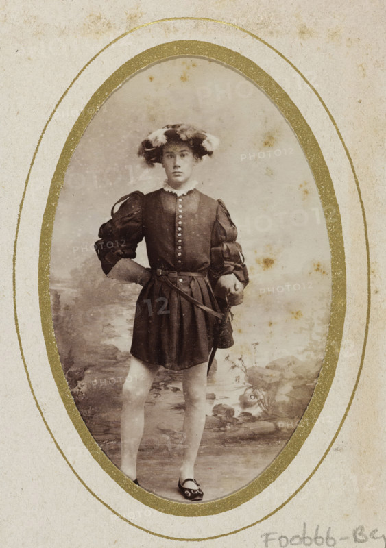 Portrait of a young man in costume with a sword, identified as M. Zijnen de Gier, album page, photograph, carte-de-visite, This photo is part of an album., name: 'M. Zijnen de Gier, 83.', inscription: 'Ant. J. van der Stok, PHOTOGRAPHE, Leiden Rapenburg 13.', photographer: Antonius Joannes van der Stok, Leiden, 1887-01-01 - 1899-12-03, paper, pasteboard, albumen print, albumendruk, height 83 mm x width 52 mm, historical persons, historical costume, hacking and thrusting weapons: rapier, An oval sepia photograph shows a young person standing in a costume with puffed sleeves and a short skirt, hands resting on hips with one hand on a belt holding a sword; the person wears a feathered hat, a high ruffled collar, and flat shoes with decorative details, and the background depicts a painted outdoor scene with rocks and foliage; the photograph is mounted on textured paper with a decorative gold oval border and some age spots visible on both the image and the mount.