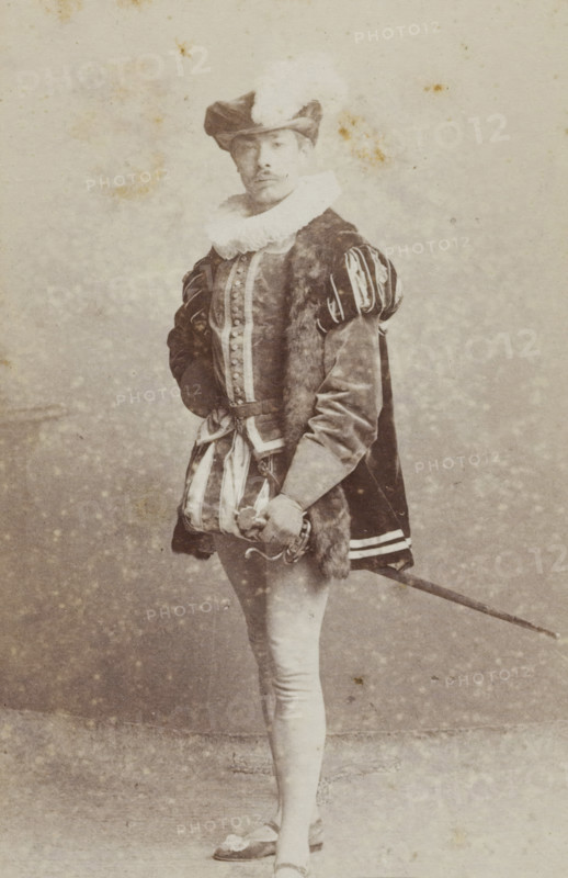 Portrait of a young man in costume with a sword, identified as NA de Joncheere, album page, photograph, carte-de-visite, This photo is part of an album., inscription: 'J. Goedeljee, Photographer, HRH PRINCE ALEXANDER, of the Netherlands, PRINCE of ORANGE, and HRH PRINCESS HENDRIK, Hoogewoerd 160, near the Plantage.', name: 'NA de Joncheere., 207.', photographer: Jan Goedeljee, Leiden, 1860 - 1905, paper, pasteboard, albumen print, height 83 mm x width 52 mm, historical persons, historical costume, hacking and thrusting weapons: rapier, A sepia-toned portrait of a person standing and facing forward, wearing a broad-brimmed hat with a turned-up side and a feather, a large ruffled collar, a fitted doublet with decorative buttons down the front, padded and slashed sleeves, a fur-trimmed cloak draped over one shoulder, striped short breeches over tight hose, flat shoes with decorative bows, and holding a sword at their side with one hand resting on the hilt.