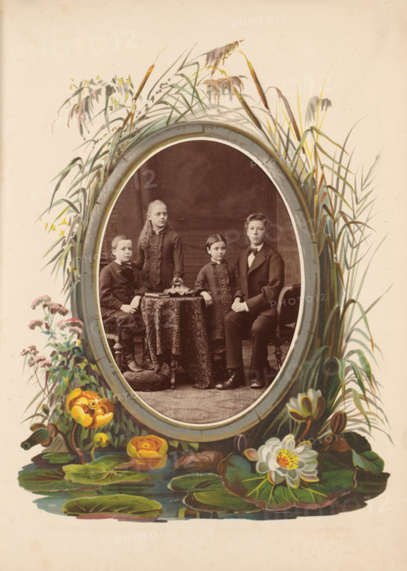 Portrait of two boys and two girls, photograph, cabinet photograph, This photo is part of an album., inscription: 'Jean Baptiste Feilner, Photograph, Bremen, Wall 185, diagonally opposite the City Theatre.', photographer: Jean Baptiste Feilner, Bremen (city), 1875 - 1899, paper, pasteboard, albumen print, height 135 mm x width 96 mm, anonymous historical persons portrayed in a group, in a group-portrait, girl (child between toddler and youth) (two persons), boy (child between toddler and youth) (two persons), An oval photograph of four children seated and standing around a small round table is centered within an illustrated floral and reed border; the children wear dark formal clothing and have neutral expressions, one boy sits on the left on a small stool, a girl stands behind the table with long hair, a younger child stands beside the table, and an older boy sits on the right on a chair; the table is covered with a patterned cloth and an object rests on top; the oval frame is surroun...