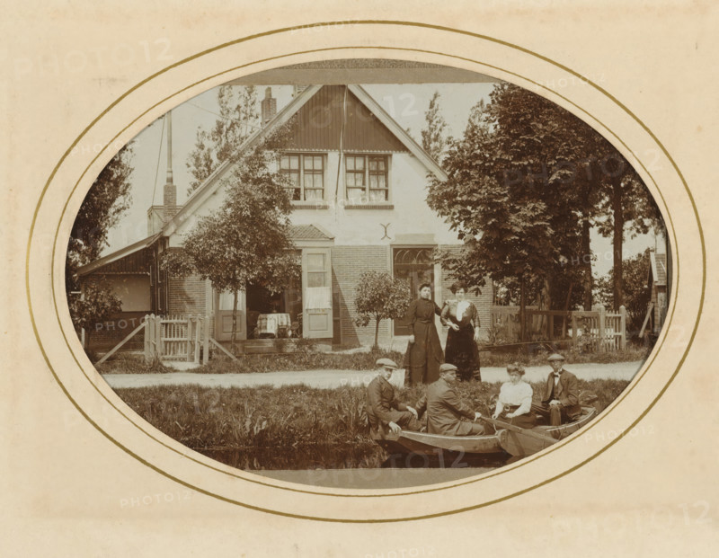 Three men and a woman in a rowboat and two women on shore, with a house in the background, album page, photograph, cabinet photograph, This photo is part of an album., inscription: 'AW van leeuwen, Photographer, Nieuwveen', photographer: AW van Leeuwen, Nieuwveen, 1883 - 1904, paper, pasteboard, daylight collodion silver print, height 137 mm x width 97 mm, rowing-boat, canoe, civic architecture, edifices, dwellings, adult woman (three persons), adult man (three persons), A sepia-toned oval photograph framed by a circular border shows a two-story house with a steep gabled roof, upper windows with curtains, a small porch with an open door, and brick and stucco exterior; trees and shrubs flank the house and a picket gate and fence run along the front; two women in long dresses stand on the lawn near the house while a group of people sit in small rowboats on a canal in front of the lawn, some wearing hats and period clothing.
