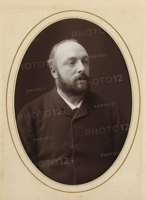 Portrait of a man, album page, photograph, cabinet photograph, This photo is part of an album., inscription: 'Inalterable Process, Charcoal Photography, Nestor Schaffers, Photographer, Rue Guinard 9, Ghent', inscription: 'Inalterable charcoal impression', photographer: Paul Joseph Nicolas Nestor Schaffers, 1875 - 1896, paper, pasteboard, carbon print, height 134 mm x width 96 mm, historical persons, adult man, moustache, A sepia-toned oval portrait of a man with a full beard and mustache visible from the chest up, set against a dark background and framed by a light-colored mat with concentric oval borders; the man wears a dark buttoned coat with a notched collar and a light shirt peeking at the neck, his hair is short and receding, and his gaze is directed slightly to one side.