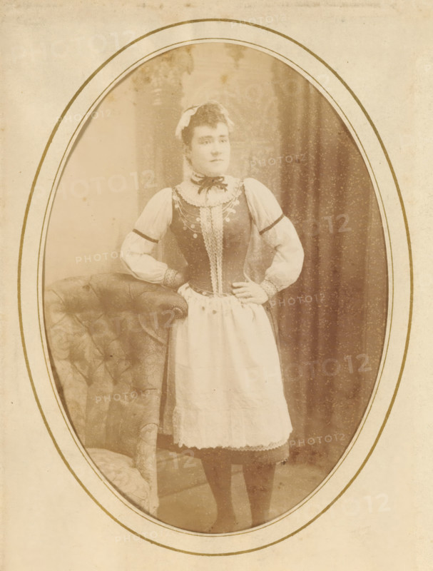 Portrait of a woman, album page, photograph, cabinet photograph, This photo is part of an album., inscription: 'Charles Thiel, ', photographer: Charles Thiel (Ainé), 1881 - 1902, paper, pasteboard, daylight collodion silver print, height 134 mm x width 96 mm, historical persons, adult woman, standing figure, A sepia oval portrait showing a woman standing with one hand on her hip and the other resting on the back of a tufted upholstered chair, wearing a dress with a fitted bodice, puffed sleeves, a lacy collar and apron, a ribbon tied at her neck, and a small headpiece, with a draped backdrop behind her and an ornamental oval mat surrounding the image.