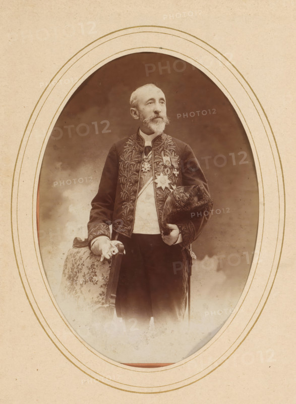 Portrait of Jules Claretie, album page, photograph, cabinet photograph, This photo is part of an album., annotation: '21407, Claretie', inscription: 'R. Dechavannes, Paris', inscription: 'Professeur Stebbing, Photographe hors concours, Boulevard des Italiens et 30, Rue de Grammont, collector's mark: 'Albert Hautecoeus, 12, Boulevard des cap..., photographer: Édouard Stebbing, Paris, 1870 - 1900, Édouard Stebbing, photographer, paper, pasteboard, albumen print, height 137 mm x width 97 mm, Person, Jules Claretie, historical persons, portrait of a writer, An oval-framed sepia photograph shows an older man standing and facing slightly to the side, wearing a dark embroidered jacket with ornate leaflike patterns and a light-colored waistcoat underneath; he has a trimmed beard and mustache and a high collar at his neck. Medals and decorative insignia are pinned to his chest. He holds a fur-trimmed hat in one hand and rests the other hand on the back of a patterned upholstered chair with a fl...
