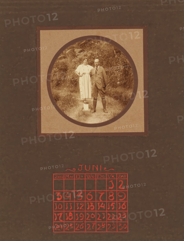 Portrait of a couple in a natural setting, album page, photograph, This photo is part of an album., annotation: 'JUNE, ', Indonesia, 1880 - in or before 1923, photographer, pasteboard, paper, daylight collodion silver print, height 78 mm x width 78 mm, historical persons, historical persons - BB - woman, marriage, married couple, 'matrimonium', A sepia photograph framed in a circular mat shows a woman and a man standing on a narrow dirt path with dense foliage behind them; the woman wears a light-colored dress with vertical stripes and buttons, has one hand on her hip and the other resting on the man's shoulder while standing on a small pedestal, and the man wears a suit with high boots and gaiters, a satchel slung across his body, and stands with his hands by his sides; the circular image is mounted on a square mat with a wide border, and below the mat a grid of hand-drawn red squares is visible against a dark background.