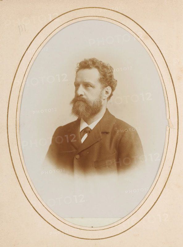 Portrait of a man with mustache and beard, album page, photograph, cabinet photograph, This photo is part of an album., inscription: 'HW Wollrabe, Hoffotograaf, The Hague, NOORDEINDE 96.', photographer: Heinrich Wilhelm Wollrabe, Dordrecht, 1880 - 1910, paper, pasteboard, daylight collodion silver print, height 137 mm x width 98 mm, historical persons, beard, mustache, A sepia-toned oval portrait of a man with wavy hair and a full beard, shown from the chest upward and facing slightly to the viewer's left, wearing a dark suit jacket with a visible lapel and a light-colored shirt with a tied neckpiece, set against a plain light background and framed by a paper mat with multiple concentric oval lines and slight edge wear.