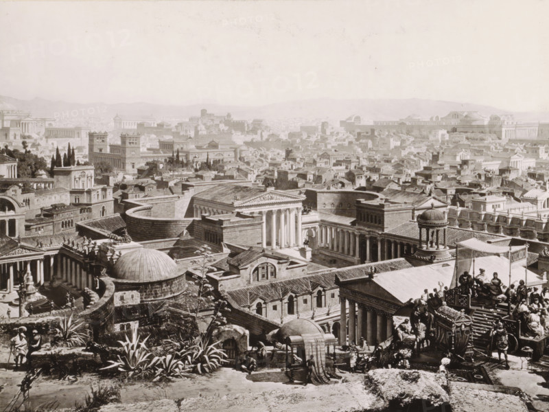 View of ancient Rome: the Forum of Augustus and the Baths of Titus, Forum des Augustus. - Baths of Titus., photograph, reproduction, page, Part of Panorama of ancient Rome., photographer: Franz Hanfstaengl, Munich, 1890, Bavaria, pasteboard, ink, materials by function, paper, printing ink, collotype, photogravure, height 179 mm x width 219 mm, prospect of city, town panorama, silhouette of city, public baths, square, circus, etc. (city(-scape) with figures, staffage), exterior, representation of a building, Rome, Forum of Augustus, Baths of Titus, A wide sepia-toned cityscape filled with densely packed classical buildings and rooftops stretching to distant hills, showing many temple-like structures with columns, domes, pediments, and colonnades, interspersed with rectangular buildings and narrow streets; in the foreground there are sculpted figures, a chariot scene with riders and attendants, plants with broad leaves, stone terraces and stairways, and a rounded structure with a domed r...