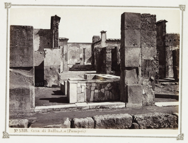 Casa di Sallustio (Pompei), Remains of the House of Sallust in Pompeii, album page, photograph, Part of Photo album with photographs of excavations in Pompeii by Giorgio Sommer., number: '5318', photographer: Giorgio Sommer, Pompeii, c. 1860 - c. 1900, paper, albumen print, height 102 mm x width 139 mm, villa, excavation, archaeology, A black and white photograph of a ruined stone structure showing several weathered walls and columns of varying textures and sizes, with rough blockwork and smoother plastered surfaces; in the foreground there is a low rectangular stone feature with a mosaic-like facing, and uneven paving and large stones lie along the bottom edge; deeper into the scene are fluted column fragments and a narrow open courtyard area framed by tall masonry blocks, shadowed recesses, and broken edges, all captured within a bordered photographic mount.