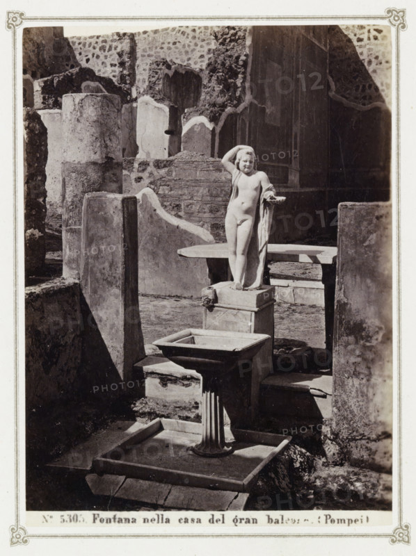 Remains of a house in Pompeii with a statue and a fountain, Fontana nella casa del gran balcone (Pompeii), album page, photograph, Part of Photo album with photographs of excavations in Pompeii by Giorgio Sommer., number: '5305', photographer: Giorgio Sommer, Pompeii, c. 1860 - c. 1900, paper, albumen print, height 140 mm x width 102 mm, piece of sculpture, reproduction of a piece of sculpture, ornamental fountain, excavation, archaeology, A stone courtyard with weathered walls and broken masonry surrounds a central sculpted fountain featuring a small nude child figure standing on a square pedestal with one hand raised to the head and a draped cloth over one arm, a shallow basin mounted on a fluted column stands in front of the pedestal, and scattered stone slabs and low platforms occupy the foreground and background.