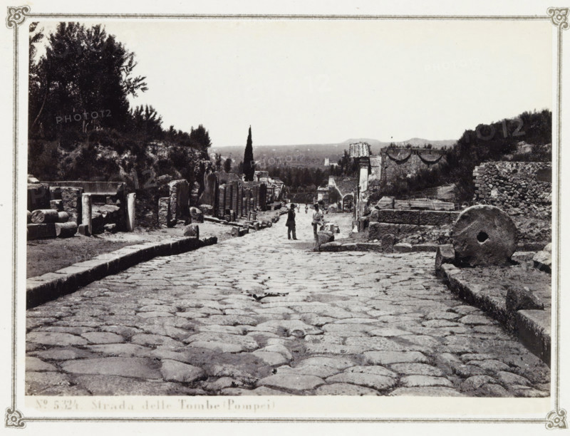 Strada delle Tombe (Pompei), Street with remains of tombs in Pompeii, album page, photograph, Part of Photo album with photographs of excavations in Pompeii by Giorgio Sommer., number: '5324', photographer: Giorgio Sommer, Pompeii, c. 1860 - c. 1900, paper, albumen print, height 103 mm x width 140 mm, street, grave, tomb, excavation, archaeology, Via delle Tombe (Pompeii), A wide cobbled street flanked by ancient stone ruins and low walls stretches toward the distance, with scattered broken columns and rectangular stone blocks along the edges, a large circular millstone resting near the right foreground, a tall slender cypress tree rising near the center background, trees and shrubs on raised embankments to the left and right, a few small human figures walking along the street, and distant low hills on the horizon.
