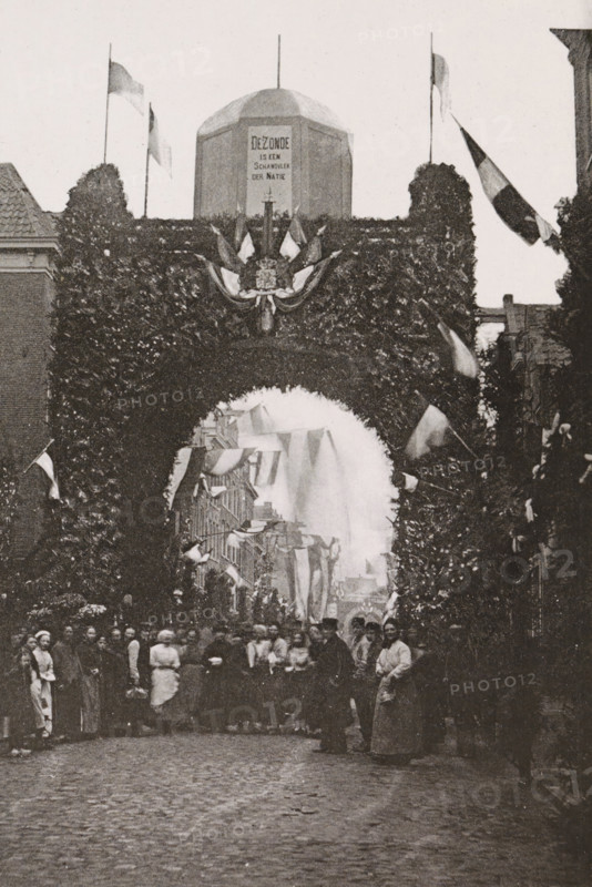 Willemstraat., Triumphal arch in the Willemstraat during the April festivities in 1887 in Amsterdam, photograph, cabinet photograph, Part of Photo album with images of the April festivities in 1887 in Amsterdam., Amsterdam, 1887-04, photographer, pasteboard, ink, materials by function, paper, printing ink, collotype, photogravure, height 147 mm x width 100 mm, prospect of city, town panorama, silhouette of city, gate, entrance, April festivities 1887, Willemstraat, A tall archway covered in dense foliage spans a cobblestone street, with a decorative crest and draped fabric at its center and flags on poles rising above; beyond the arch a narrower street is lined with buildings and more hanging banners, and a crowd of people stands beneath and in front of the arch, with some figures in long garments and others in darker coats, their faces and postures visible but not detailed.