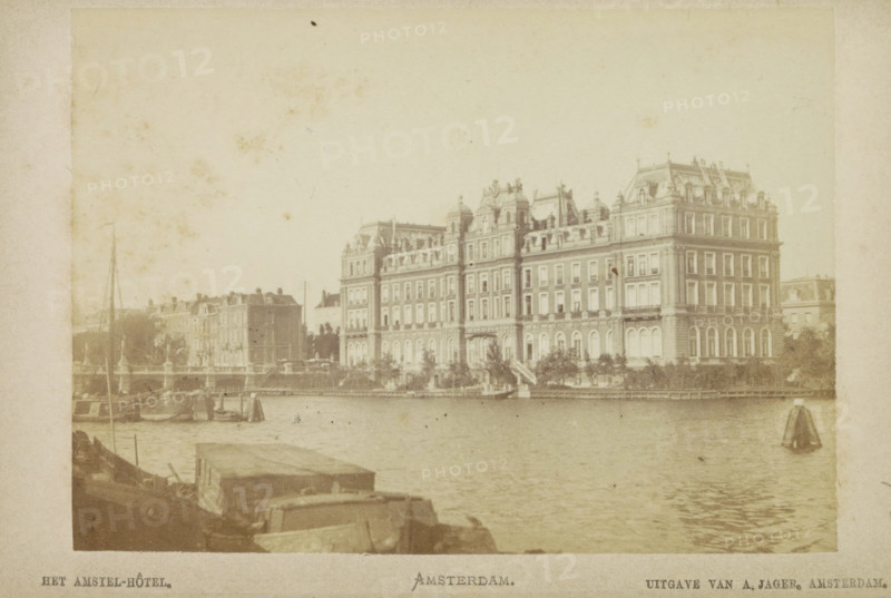 The Amstel Hotel. Amsterdam., View of the Amstel Hotel, photograph, cabinet photograph, Part of a Leporello album with 12 photographs of Amsterdam., photographer: attributed to Andries Jager, Amsterdam, c. 1860 - c. 1900, Attributed to, paper, pasteboard, albumen print, height 95 mm x width 135 mm, river, hotel, hostelry, inn, Amstel (river), Amstel Hotel, Professor Tulpplein, A waterfront scene shows a large multi-story building with ornate rooflines and many windows stretching along the shore, with steps and trees at its base; smaller buildings and architectural elements extend to the left; water occupies the foreground with a few boats and small sails visible near the shore; the image has a muted, aged tone and a pale border surrounding it.