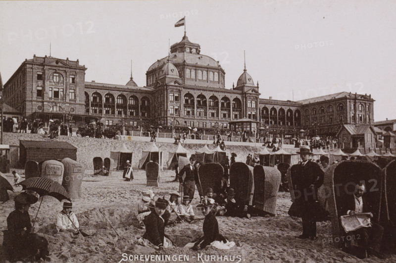 View of the Kurhaus in Scheveningen with bathers in the foreground, Scheveningen - Kurhaus., photograph, cabinet photograph, Part of a Leporello album with photographs of Scheveningen., Scheveningen, c. 1890 - c. 1910, photographer, pasteboard, ink, materials by function, paper, printing ink, collotype, photogravure, height 97 mm x width 147 mm, beach (as place of recreation), watering-place, spa, Kurhaus (Scheveningen), A wide beachfront scene shows a large ornate building with multiple arched windows, domed roof elements, and a flag atop a central cupola, sitting behind a raised promenade lined with a row of tents and a balustrade; the sandy foreground is filled with many wicker beach chairs, groups of people sitting and standing, some holding parasols, and several men in suits and hats, with one man seated reading a paper inside a chair-like shelter.