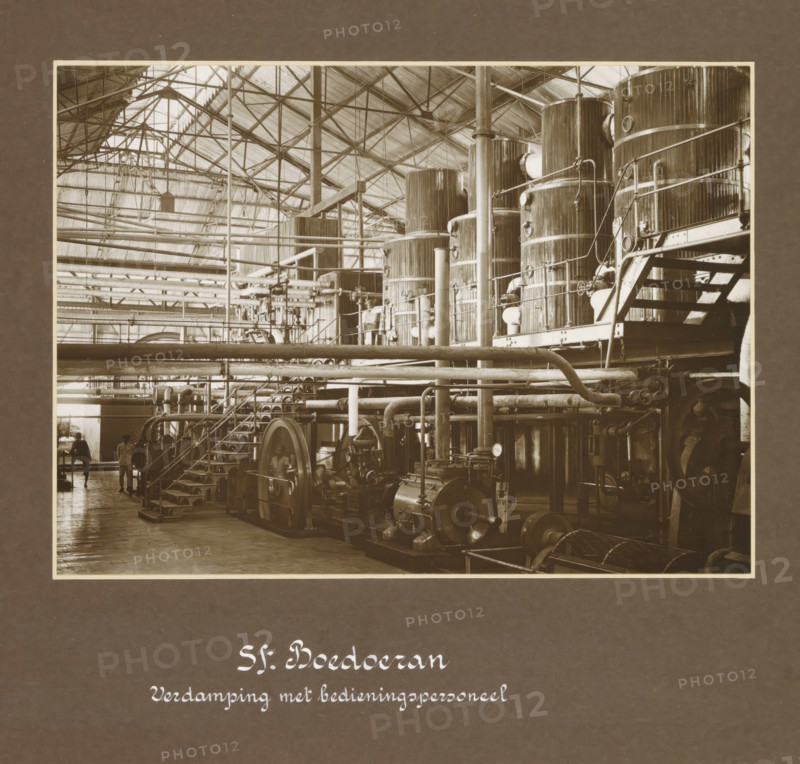 Hall with evaporation installation of the Boedoeran sugar factory in Sidoarjo on Java, Sf. Boedoeran Evaporation with operating personnel, album page, photograph, Part of Photo album with photographs of four sugar factories on Java., photographer: Isken, Sidoarjo, c. 1925 - c. 1930, Java, East Java, pasteboard, paper, gelatin silver print, developing gelatin silver print, height 297 mm x width 450 mm, plantation, sugar, factory-interior, machine shop, plants and herbs: sugarcane, Dutch East Indies, The, A large industrial interior filled with tall cylindrical tanks and vertical pipes connected by horizontal piping and metal railings, with platforms and staircases alongside the tanks; rows of beams and trusses supporting a pitched, corrugated roof overhead; heavy machinery including motors and flywheels mounted on a floor with visible piping and gauges; structural columns rising between equipment; a few figures standing near the far wall beneath the overhead pipes; tiled or wooden floor...