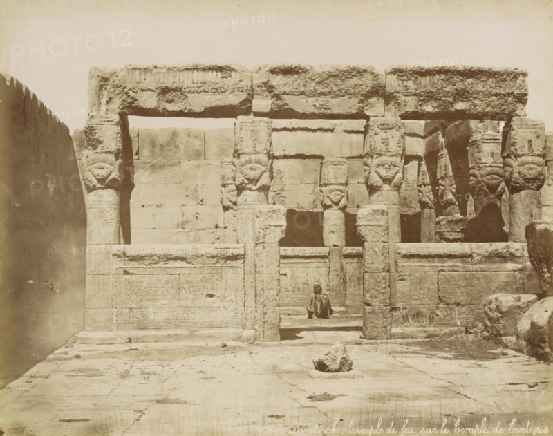 Dendérah, Temple de feu sur le temple de Tentyris, Temple complex of Dendera, photograph, Part of Travel album with 80 photos of Egypt., negative number: '125', photographer: Maison Bonfils, Dendera, 1879, North Africa, pasteboard, paper, albumen print, height 220 mm x width 279 mm, temple, shrine, Egyptian religion, A large stone facade of an ancient structure fills the frame, showing worn columns and lintels carved with decorative reliefs and symbols, some tops of columns shaped with stylized heads. The stone surfaces display weathering and textured erosion, with rectangular blocks forming the wall behind the columns. A person sits on a step within a recessed doorway in the center of the composition, wearing layered garments and a head covering. The foreground shows a paved stone courtyard with a rough rock placed near the middle and scattered debris on the ground.