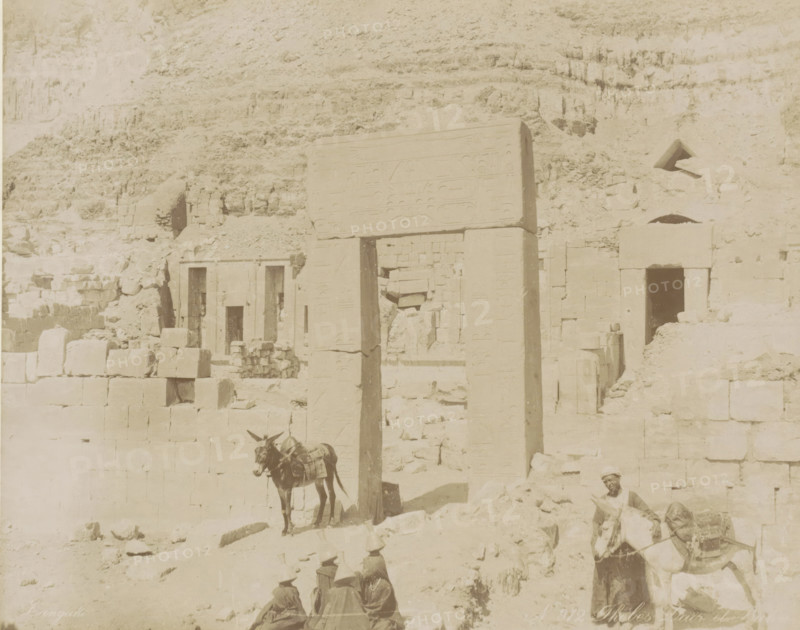 People and donkeys at the temple complex of Deir el-Bahri, Thebes Dair el Ba, photograph, Part of Travel album with 80 photos of Egypt., negative number: '912', photographer: Zangaki, C. & G., Deir el-Bahri, c. 1870 - c. 1890, North Africa, pasteboard, paper, albumen print, height 216 mm x width 275 mm, temple, shrine, Egyptian religion, A sunlit scene of weathered stone ruins set against a rocky cliff, showing a tall carved stone gateway with vertical columns and a horizontal lintel bearing shallow reliefs, adjacent stone blocks and partially intact walls with rectangular doorways and small openings, rubble and scattered stones on the uneven ground, a donkey standing near the gateway with packs on its back, a man holding another donkey near the right edge wearing light clothing and a head covering, and three seated figures wearing conical hats in the foreground.