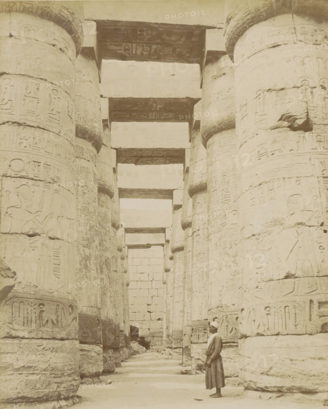 Temple of Karnak, Thebes (Luxor), Man in a colonnade with hieroglyphs (former title), photograph, Part of Travel album with 80 photos of Egypt., negative number: '1011', photographer: Zangaki, C. & G., Egypt, c. 1870 - c. 1890, North Africa, pasteboard, paper, albumen print, height 275 mm x width 220 mm, colonnade, Egyptian hieroglyphs, Luxor, A sepia-toned photograph showing a row of large, cylindrical stone columns with carved reliefs and rounded bands, forming a colonnade that recedes into the distance beneath horizontal stone lintels, with a tall stone wall visible at the far end and a single man in traditional clothing and a head covering standing barefoot near the foreground looking toward the columns.