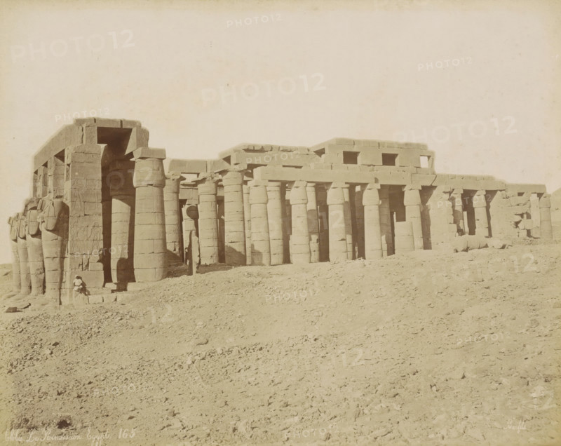 Thebes Le Ramesseum Egypt, Exterior of the Ramesseum in Thebes (Luxor), photograph, Part of Travel Album with 80 Photos of Egypt., negative number: '165', photographer: Maison Bonfils, Thebes, c. 1870 - c. 1898, North Africa, Upper Egypt, pasteboard, paper, albumen print, height 221 mm x width 280 mm, temple, shrine, Egyptian religion, A wide view of an ancient stone structure with a colonnade of tall, rounded columns supporting horizontal lintels, several carved statues and reliefs visible on the leftmost pillars, a large entrance framed by massive blocks, rubble and sandy ground in the foreground, and a lone seated figure near the base of the leftmost statues.