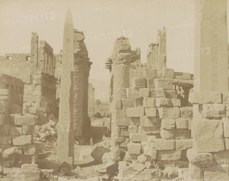 Karnak Grand temple with obélisques de Touthmes 1er et de sa fille Hatasou, Great Temple with the obelisks of Thutmose I and his daughter Hatshepsut in Karnak, photograph, Part of Travel album with 80 photos of Egypt., negative number: '994', photographer: Zangaki, C. & G., Luxor, c. 1870 - c. 1890, North Africa, pasteboard, paper, albumen print, height 216 mm x width 273 mm, obelisk, needle, temple, shrine, Egyptian religion, A wide view of an ancient stone ruin with tall carved columns and an obelisk standing among stacks of large rectangular blocks, weathered surfaces and broken masonry visible throughout, a narrow passageway between the structures leading to a distant palm tree, a few small human figures near the base of the ruins, and the scene rendered in a muted, sepia tone.
