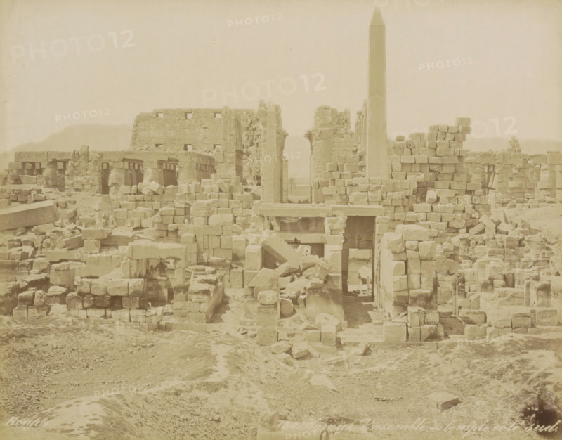 South side of the Karnak temple complex, Karnak Ensemble du temple, côté sud, photograph, Part of Travel Album with 80 Photos of Egypt., negative number: '150', photographer: Maison Bonfils, Luxor, c. 1870 - c. 1898, North Africa, pasteboard, paper, albumen print, height 219 mm x width 282 mm, temple, shrine, Egyptian religion, A wide view of weathered stone ruins with scattered blocks and collapsed walls across a dusty foreground, rectangular stone lintels and doorways visible among stacked masonry, a tall obelisk rising centrally above the rubble, fractured columns and carved stone elements interspersed with rubble piles, a large partially intact wall in the background and low hills on the horizon, overall tones are pale and monochromatic.