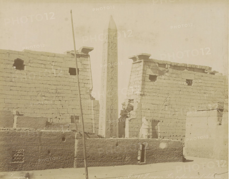 Louqsor Le pylon avec l'obelisque, Pylon with obelisk in Luxor, photograph, Part of Travel album with 80 photos of Egypt., negative number: '971', photographer: Zangaki, C. & G., Luxor, c. 1870 - c. 1890, Zangaki, photographer, North Africa, pasteboard, paper, albumen print, height 217 mm x width 276 mm, obelisk, needle, gate, entrance, A sepia-toned photograph shows a tall obelisk with carved vertical panels rising between two ancient stone walls topped with flat cornices, the walls marked by rectangular openings and surface carvings. A vertical pole leans in front of the left wall and a fragment of a statue stands near an entrance at the base of the right wall. The foreground includes a lower textured masonry wall with a small arched doorway and geometric lattice opening, and scattered stones and rubble rest along the base of the structures. The sky above is pale and empty.