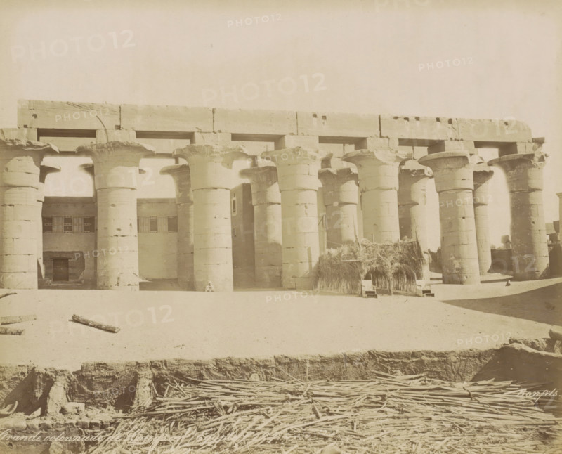 Grande colonnade de Louqsor (Égypte), Temple of Luxor, Great colonnade in Luxor (former title), photograph, Part of Travel Album with 80 Photos of Egypt., negative number: '127', photographer: Maison Bonfils, Luxor, c. 1870 - c. 1898, North Africa, pasteboard, paper, albumen print, height 224 mm x width 280 mm, colonnade, A row of large stone columns with flared capitals supports a heavy horizontal lintel, forming an open colonnade in front of a stone building with rectangular window openings; a small person sits at the base of one column; bundles of reeds or thatch are stacked against part of the colonnade near a simple woven shelter; scattered logs and sticks lie on the sandy ground in the foreground, where an exposed trench edge reveals masonry and piled wooden poles below.