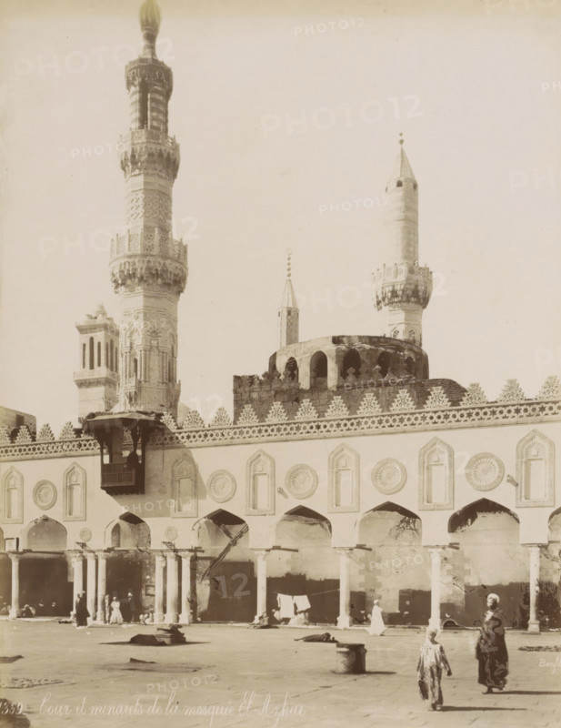 Courtyard and minarets of the Al-Azhar Mosque in Cairo, Cour et minarets de la mosquée El-Azhar, photograph, Part of Travel Album with 80 Photos of Egypt., negative number: '1369', photographer: Maison Bonfils, Cairo (city), c. 1870 - c. 1898, Ottoman Empire, North Africa, pasteboard, paper, albumen print, height 280 mm x width 218 mm, temple, shrine, Islam, Mohammedanism, An exterior view of a large ornate building with arched openings along its facade and decorative circular and pointed panel designs above each arch, a patterned crenellated parapet along the roofline, and tall slender towers rising behind it with carved and geometric detailing on their shafts and ornate balconies, one tower topped with a bulbous finial and another with a conical top and small spherical ornaments above it; a wooden projecting balcony with carved supports is attached to the facade, a rounded domed section with arched openings sits behind the parapet, and stone columns support the arcade below where a f...