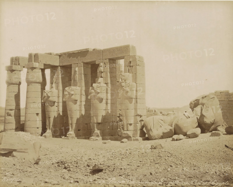 Overturned Statue of Sesostris in Thebes (Egypt), Overturned statue of Pharaoh Senusret (Sesostris) in Thebes (former title), Ramesseum (Temple of Ramses II), Thebes (Luxor), photograph, Part of Travel Album with 80 Photos of Egypt., negative number: '167', photographer: Maison Bonfils, c. 1870 - c. 1898, North Africa, Upper Egypt, pasteboard, paper, albumen print, height 221 mm x width 281 mm, monument, statue, Luxor, A row of tall stone columns and statues stands on a sandy, rocky ground, with several columns topped by square capitals and adjacent statues carved in a seated or standing pose, some of the statues appearing damaged or fragmented; a large broken statue with a visible head lies among scattered stone blocks and rubble to the right, while additional fallen fragments and stones are scattered across the foreground; a low stone wall or structure is visible behind the fallen pieces at the far right, and the sky above is clear and pale.