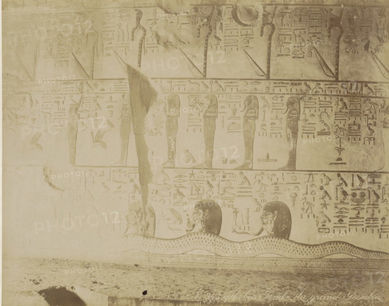 Tomb of Seti I (KV17) in the Valley of the Kings, Thebes (Luxor), Burial chamber of pharaoh Senuseret (Sesostris) in Thebes (former title), Thebes Tombe du grand Sesotris, photograph, Part of Travel album with 80 photos of Egypt., negative number: '943', photographer: Zangaki, C. & G., c. 1870 - c. 1890, North Africa, Upper Egypt, pasteboard, paper, albumen print, height 218 mm x width 276 mm, grave, tomb, Egyptian hieroglyphs, Luxor, A faded wall panel with carved figures and symbols in a monochrome sepia tone, showing vertical standing humanoid figures with headdresses aligned along horizontal bands, smaller bird and animal shapes interspersed among rectangular and linear carved motifs, repeating rows of hieroglyph-like pictorial signs, a large undulating serpent-like form along the lower band with patterned scales, partial human heads emerging above the serpent with visible facial features and hair, areas of surface wear and patched plaster with lighter and darker discolorations, an...