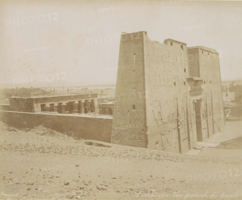 Edfou Vue general e du temple, View of the Temple of Horus in Edfu, photograph, Part of Travel album with 80 photos of Egypt., negative number: '1025', photographer: Zangaki, C. & G., Temple of Horus (Edfu), c. 1870 - c. 1890, North Africa, pasteboard, paper, albumen print, height 219 mm x width 268 mm, temple, shrine, Egyptian religion, A large stone structure with tall, sloping walls covered in carved figures occupies the right side of the image, showing doorways and reliefs and a small projecting balcony, while to the left a row of columns and a low surrounding wall sit behind a sandy foreground scattered with small stones and depressions, and in the distant background there is a flat landscape with low vegetation and a few taller trees under a pale sky.