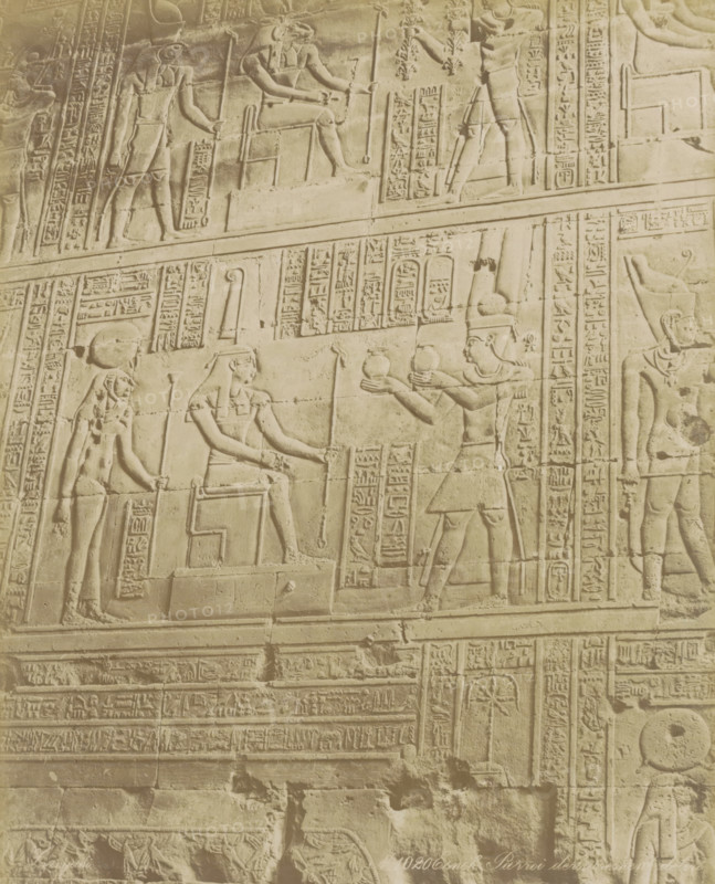 Esneh Parroi der, de te, Wall with hieroglyphs and reliefs in Esna, photograph, Part of Travel album with 80 photos of Egypt., negative number: '1020', photographer: Zangaki, C. & G., Esna, c. 1870 - c. 1890, North Africa, pasteboard, paper, albumen print, height 272 mm x width 220 mm, sculpture (relief, sculpture), Egyptian hieroglyphs, A carved stone wall panel showing raised relief figures and decorative bands, with multiple humanlike figures in profile wearing headdresses and holding staffs or offering vessels, seated and standing poses visible, framed by vertical and horizontal panels filled with repeated pictorial symbols, the surface showing signs of wear and faint discoloration, and rows of ornamental carved borders separating different registers of the scene.