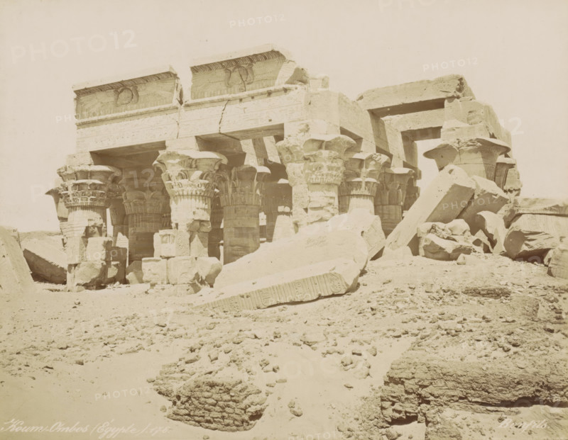Koum-Ombos (Égypt), View of the Temple of Kom Ombo, photograph, Part of Travel album with 80 photos of Egypt., number: '89', negative number: '176', photographer: Maison Bonfils, Kom Ombo, c. 1870 - c. 1898, North Africa, pasteboard, paper, albumen print, height 220 mm x width 280 mm, temple, shrine, Egyptian religion, A sunlit ruin of a stone structure with rows of large carved columns topped by ornate capitals, some capitals shaped like stylized plant forms, stands amid scattered massive stone blocks and broken architectural fragments lying on a sandy, rubble-strewn ground, with horizontal lintels and a partially intact entablature showing carved reliefs above the columns and open spaces between the supports.