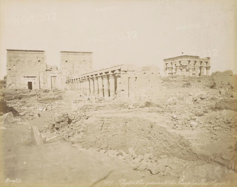 Phyloe. General view of the temples of Isis (Egypt), View of the Temples of Isis on the island of Philae, photograph, Part of a travel album with 80 photos of Egypt., negative number: '187', photographer: Maison Bonfils, Philae, c. 1870 - c. 1898, North Africa, pasteboard, paper, albumen print, height 222 mm x width 280 mm, The Temples of Isis on Philae were moved to the island of Agilkia between 1977 and 1980 due to the construction of the Aswan Dam., temple, shrine, Egyptian religion, Egypt, A wide, pale photograph of an ancient stone temple complex showing large, flat-topped walls and gateways with carved reliefs, a long colonnade of round columns with decorated capitals stretching diagonally across the middle ground, and a separate pavilion with clustered columns on the right, all set amid a foreground of uneven, rocky ground and scattered stone blocks.