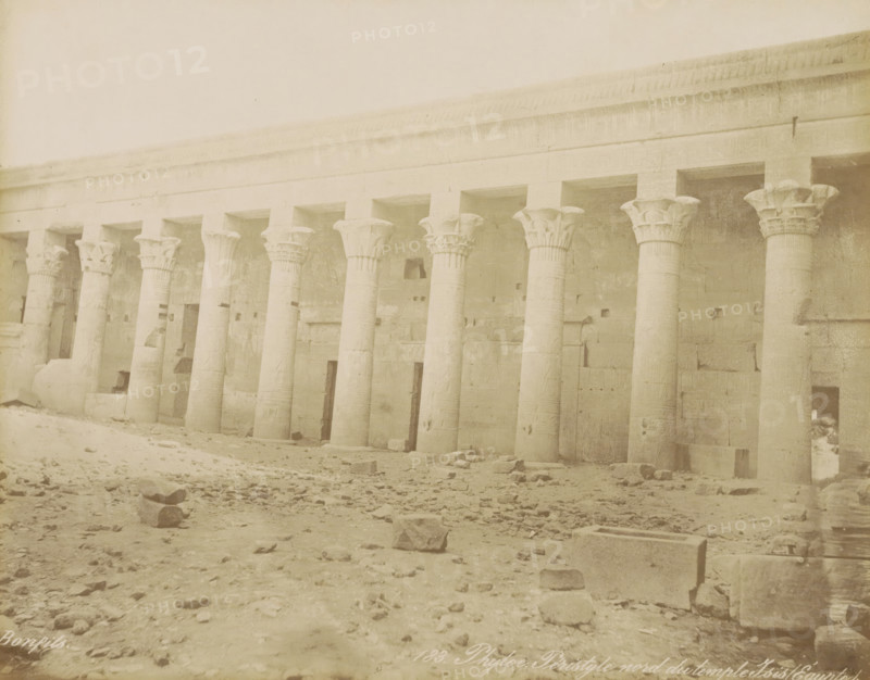 Phyloe. Peristyle nord du temple Isis (Egypt), Northern colonnade (peristilium) of the Temple of Isis on the island of Philae, photograph, Part of Travel album with 80 photos of Egypt., negative number: '183', photographer: Maison Bonfils, Philae, c. 1870 - c. 1898, North Africa, pasteboard, paper, albumen print, height 221 mm x width 280 mm, colonnade, temple, shrine, Egyptian religion, Temple of Isis (Philae), A wide exterior view of a long stone colonnade with tall carved columns topped by ornate capitals, each column set into a recessed bay beneath a continuous entablature, a rough stone wall behind the columns showing shallow niches and rectangular openings, a sandy and rubble-strewn foreground with scattered blocks and debris, and a pale sky above.