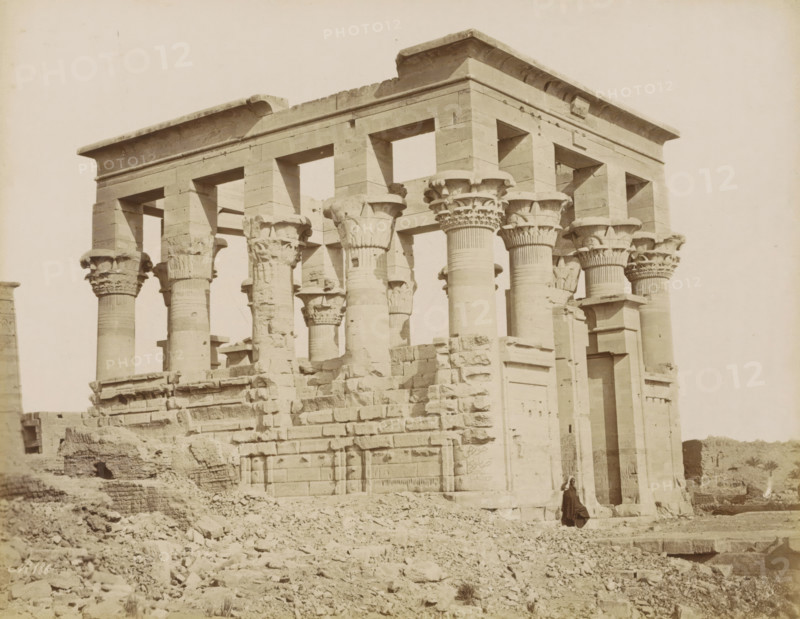 Temple on the island of Philae, photograph, Part of Travel album with 80 photos of Egypt., negative number: '186', photographer: attributed to Zangaki, C. & G., Philae, c. 1870 - c. 1890, North Africa, Attributed to, pasteboard, paper, albumen print, height 219 mm x width 280 mm, colonnade, temple, shrine, Egyptian religion, A weathered stone temple façade with tall fluted columns topped by ornate capitals, a horizontal entablature with rectangular openings above the columns, and a rubble-strewn foreground; portions of the stonework show chips and erosion, and a solitary figure in dark clothing stands near a doorway at the base of the structure.