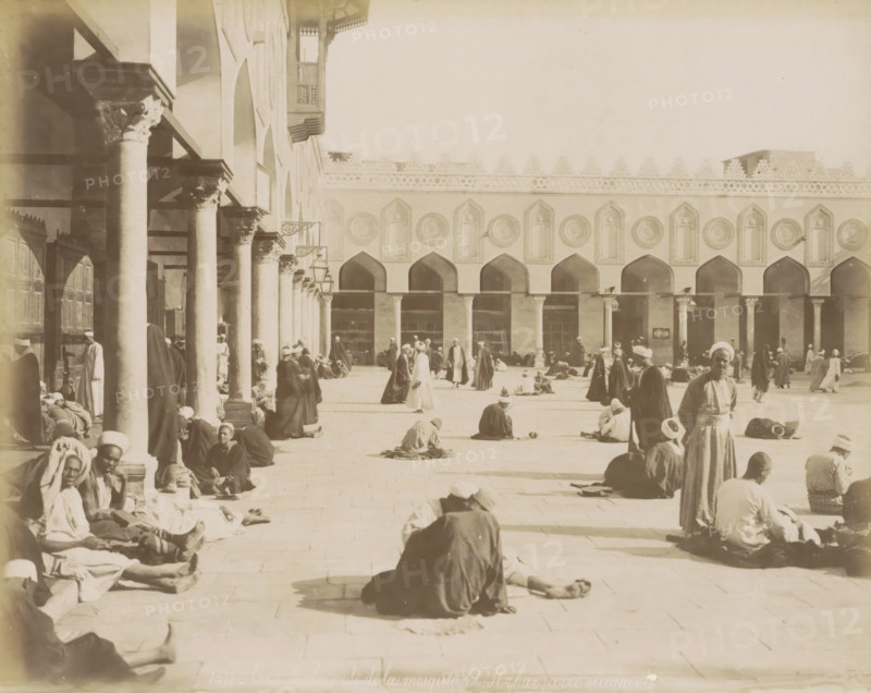 Students in the courtyard of the Al-Azhar Mosque in Cairo, Courtyard and colonnade of the Al-Azhar Mosque, with students, photograph, Part of a travel album with 80 photos of Egypt., negative number: '1371', photographer: Maison Bonfils, Cairo (city), c. 1870 - c. 1898, Ottoman Empire, North Africa, pasteboard, paper, albumen print, height 224 mm x width 280 mm, temple, shrine, Islam, Mohammedanism, A wide sunlit courtyard bordered by a building with a row of tall arches and decorative round medallions above each arch, and a colonnaded portico with columns and carved capitals along one side; many people wearing long robes and head coverings sit, stand, walk, or recline across the paved ground, some grouped near the columns and others scattered toward the center and far side; shadows from the columns fall across the courtyard; a few hanging lamps and ornate bracketed fixtures are attached to the portico; architectural details include carved patterns along the upper wall and a projecting...