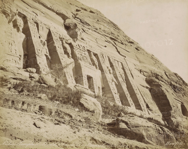 Nubie. Temple d' Abou Simbel, dédie a Athor (Égypt), Temple of Abu Simbel, photograph, Part of Travel album with 80 photos of Egypt., negative number: '201', photographer: Maison Bonfils, Abu Simbel, c. 1870 - c. 1898, Maison Bonfils, photographer, North Africa, Upper Egypt, pasteboard, paper, albumen print, height 221 mm x width 279 mm, sculpture (relief, sculpture), temple, shrine, Egyptian religion, A steep rock face with carved vertical recesses and sculpted humanlike figures integrated into the stone, fluted columns and rectangular doorways set into the cliff, patterned reliefs decorating the carved surfaces, scattered boulders and ledges at the base, sparse dry vegetation growing among the rocks, and a pale sky above.