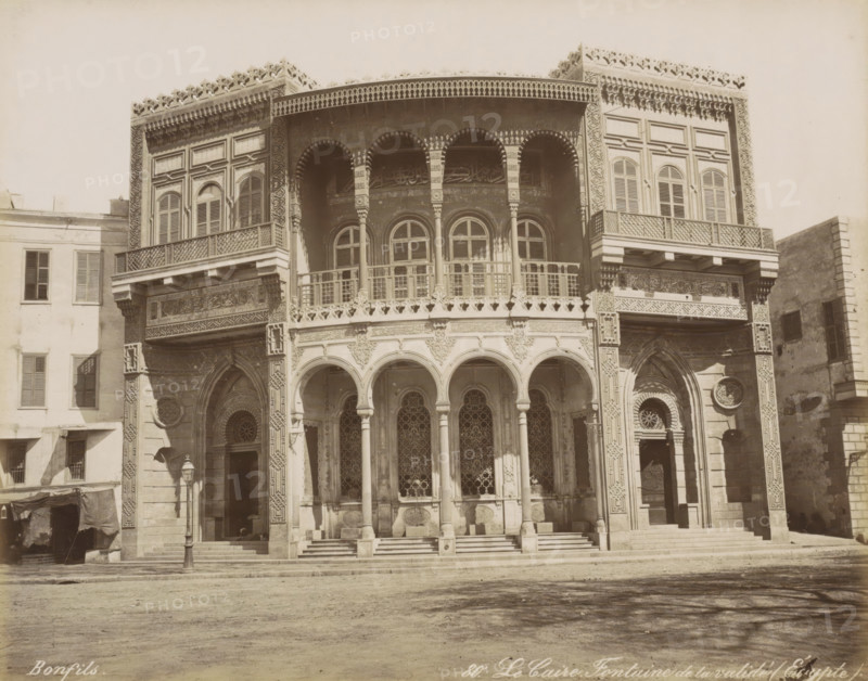 Le Caire, Fontaine de la Validé (Egypt), Building with well in Cairo, photograph, Part of Travel Album with 80 Photos of Egypt., negative number: '80', photographer: Maison Bonfils, Cairo (city), c. 1870 - c. 1898, Ottoman Empire, North Africa, pasteboard, paper, albumen print, height 220 mm x width 281 mm, utilitarian, public pump or water fountain, A frontal view of a large ornate building facade with an upper level featuring rows of arched windows and decorative railings, and a lower level with a series of tall arches supported by slender columns leading to intricately patterned window grilles and doorways; the facade displays carved and geometric ornamentation around arches and windows, latticework balconies, and decorative friezes along the top edge; steps lead up to the entrance area and a street lamp stands near one side; adjacent buildings with shuttered windows flank the ornate structure and a bare ground surface occupies the foreground.