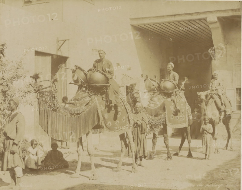 the Arab marriage, Wedding procession with camels in Egypt, photograph, Part of Travel album with 80 photos of Egypt., photographer: Zangaki, C. & G., Egypt, c. 1870 - c. 1890, North Africa, pasteboard, paper, albumen print, height 218 mm x width 275 mm, members of bridal party, A sunlit sepia photograph shows decorated camels with ornate blankets, fringes, and hanging adornments, ridden by men wearing turbans and robes; some riders sit atop large drums mounted on the camels while other men and children stand or sit nearby in traditional garments, with a building featuring a shaded archway and a doorway forming the background.