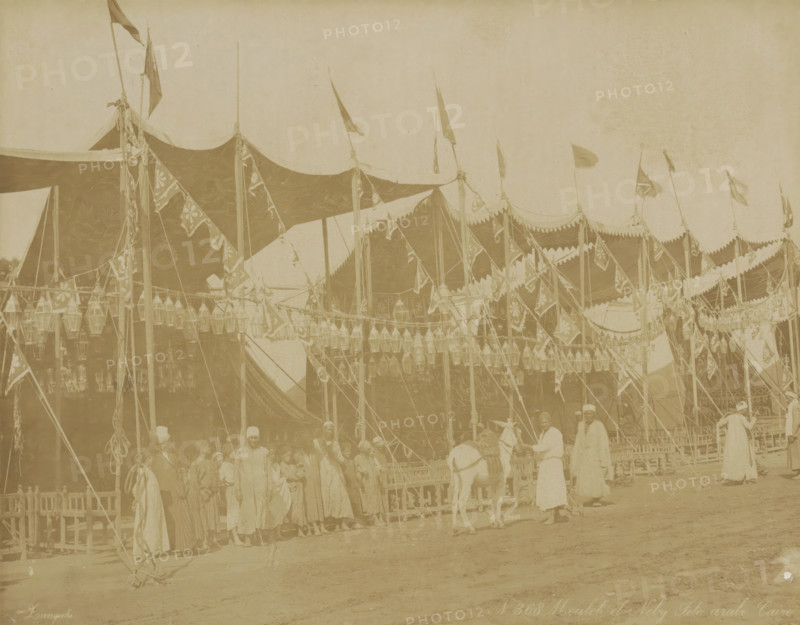 Decorated festival grounds in Cairo, Moulet el Neby Fête Arabe Caire, photograph, Part of Travel Album with 80 Photos of Egypt., negative number: '368', photographer: Zangaki, C. & G., Cairo (city), c. 1870 - c. 1890, Ottoman Empire, North Africa, pasteboard, paper, albumen print, height 219 mm x width 276 mm, festivities, A large striped tent with scalloped edges and decorative hanging banners and lanterns stretches across the scene, supported by tall poles topped with fluttering flags; a group of people in long robes and head coverings stand and gather near the tent while a person leads a donkey in front; wooden fencing and ropes are visible around the area, and the ground appears dusty.