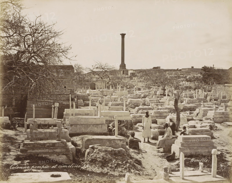 Cemetery with column in Egypt, Colonne pompée et cimetière, photograph, Part of Travel album with 80 photos of Egypt., negative number: '22', photographer: Maison Bonfils, Egypt, c. 1870 - c. 1898, North Africa, pasteboard, paper, albumen print, height 223 mm x width 281 mm, churchyard, cemetery, A sepia-toned scene of a graveyard with rows of stone tombs and raised rectangular grave markers spread across uneven ground, scattered bare trees with twisted branches, a tall solitary column rising in the distant background from a stone base, low stone walls and buildings along the far edge of the burial area, a group of people in simple garments gathered among the graves with some seated on stone ledges and one standing, stone pathways running between the tombs, a few plants and agave-like leaves near some graves, and a wooden gate or fence segment visible near a stone wall on the left.