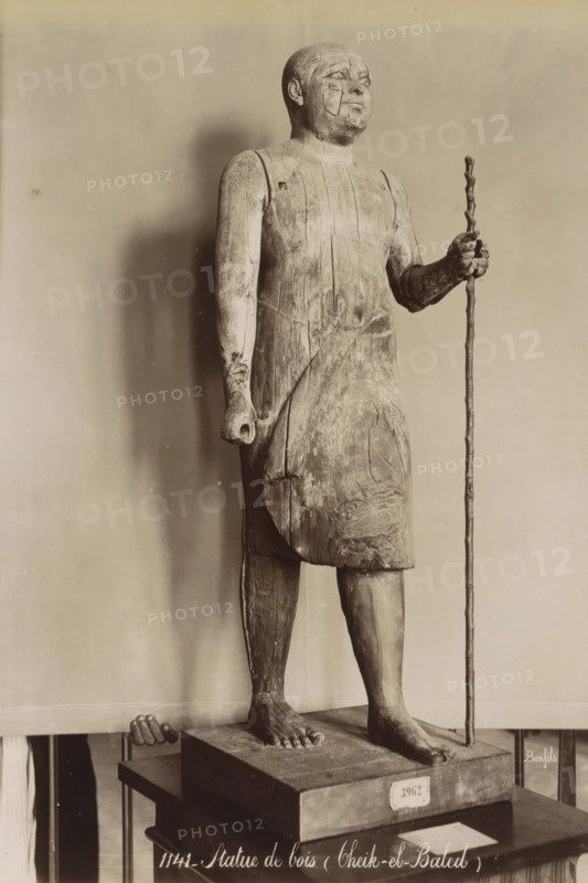 Wooden statue of a man with a staff (Cheik-el-Baled), Statue de bois (Cheik-el-Baled), photograph, Part of Travel Album with 80 Photos of Egypt., negative number: '1141', photographer: Maison Bonfils, Egypt, c. 1870 - c. 1898, North Africa, pasteboard, paper, albumen print, height 279 mm x width 216 mm, monument, statue, A full-length wooden figure stands on a rectangular base, shown from head to feet and holding a tall, thin staff in one hand; the figure wears a simple sleeveless tunic, has short hair, and a weathered surface with visible wood grain and cracks; one arm hangs by the side with a closed hand while the other is bent to grip the staff, and the bare feet rest flat on the base; a small hand belonging to a person is visible at the left edge near the base; the background is plain and evenly lit.