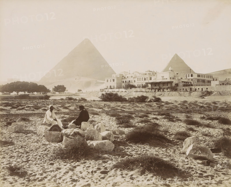 Hotel Mena - House and pyramids, View of the Pyramids of Giza with Hotel 'Mena House' in the foreground, photograph, Part of Travel Album with 80 Photos of Egypt., negative number: '1434', photographer: Maison Bonfils, Giza, c. 1886 - c. 1898, North Africa, pasteboard, paper, albumen print, height 221 mm x width 272 mm, hotel, hostelry, inn, pyramid (historical grave form), A wide sandy landscape with scattered low shrubs and rocks in the foreground shows two seated figures on a cluster of stones, one wearing a light robe and head covering and the other in darker clothing and a brimmed hat, facing each other. Behind them a built cluster of multi level stone and stucco buildings with flat roofs and a small domed structure runs along a raised embankment, with trees visible to the side. Two large triangular stone pyramids rise in the background behind the buildings, occupying much of the sky area. The overall scene is sepia toned with textured sand and sparse vegetation across the foregro...